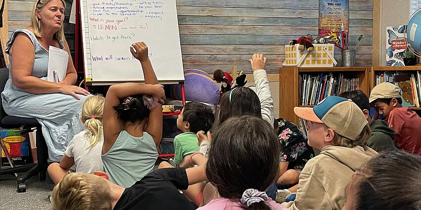 Teacher sitting next to an easel.  Students sitting on the floor in front of the easel.  Several students are raising their han