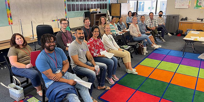 15 adults sitting in a classroom. Rainbow rug in front of them. 