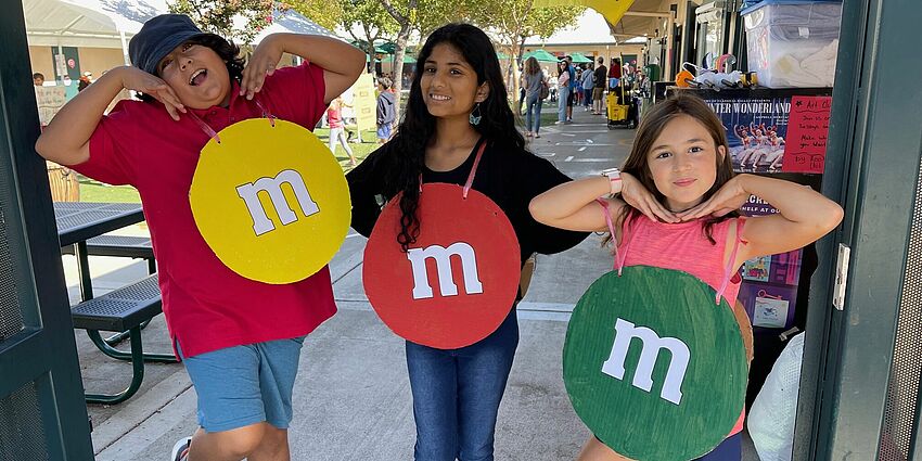 Three kids with yellow, red and green M&M costumes on. Smiling in gateway to school. Yellow flag flying behind them.