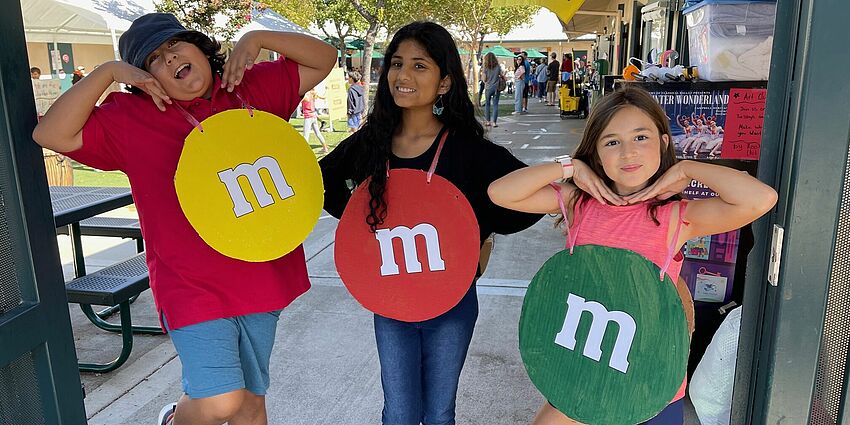 Three kids with yellow, red and green M&M costumes on.  Smiling in gateway to school.  Yellow flag flying behind them.  