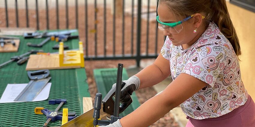 Girl wearing pink is sawing a piece of wood while wearing safety glasses. 