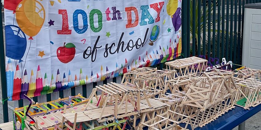 Several popsicle-stick bridges lined up on a table in front of banner that says 100th Day of School