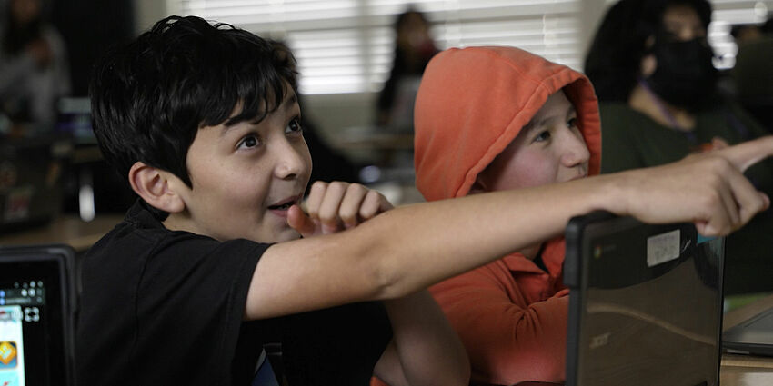 middle school boy at desk looks excited as he points in front of him.