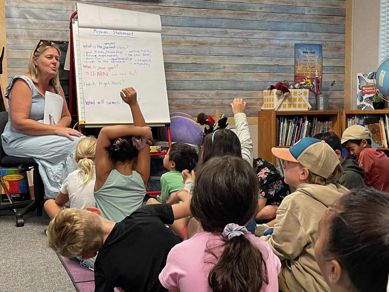 Teacher sitting next to an easel.  Students sitting on the floor in front of the easel.  Several students are raising their han