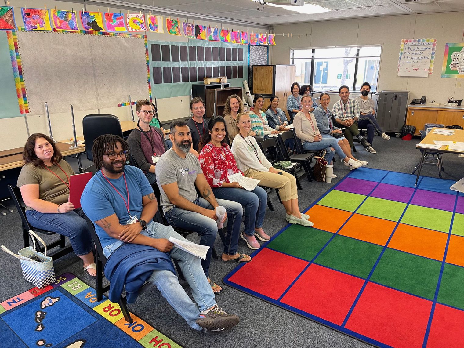 15 adults sitting in a classroom. Rainbow rug in front of them. 