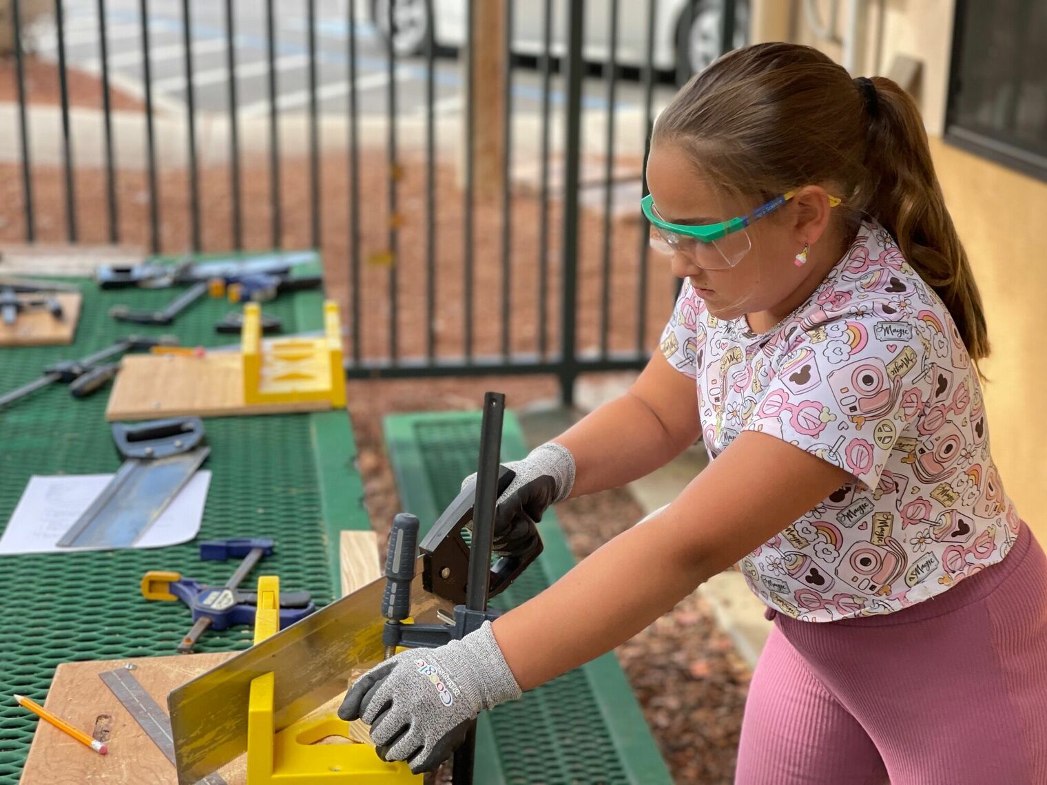 Girl wearing pink is sawing a piece of wood while wearing safety glasses. 