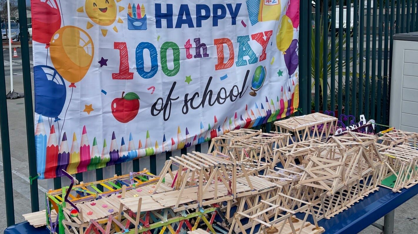 Several popsicle-stick bridges lined up on a table in front of banner that says 100th Day of School